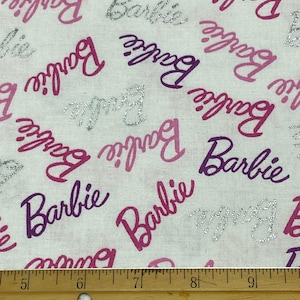 May include: White fabric with a repeating pattern of the word "Barbie" in various shades of pink and silver glitter. A wooden ruler is at the bottom of the image, with measurements in inches.