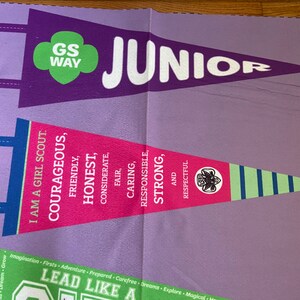 May include: A purple and green pennant with the text "GS WAY JUNIOR" and the words "I am a Girl Scout. Courageous, Friendly, Honest, Considerate, Fair, Caring, Responsible, Strong, and Respectful."