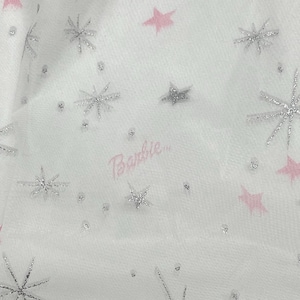 May include: White fabric with a pattern of silver glitter snowflakes, pink stars, and silver glitter dots. The word "Barbie" is written in pink script. The fabric has a sheer quality.