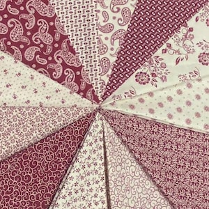 May include: A circular arrangement of fabric wedges in shades of burgundy and cream. The wedges display various patterns, including paisley, floral, and small dot designs. The fabrics are arranged in a radial pattern, creating a visually appealing textile design.