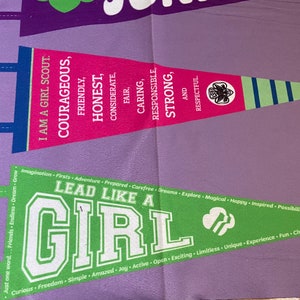 May include: A green and white pennant with the text "Lead Like A Girl" and the Girl Scout logo. The pennant is hanging on a purple background with a pink pennant above it. The pink pennant has the text "I am a Girl Scout" and lists the Girl Scout values: Courageous, Friendly, Honest, Considerate, Fair, Caring, Responsible, Strong, and Respectful.