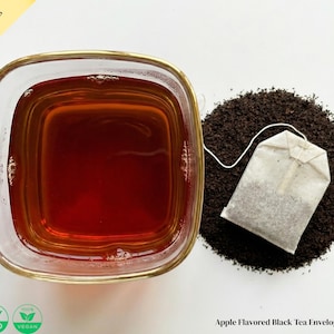 May include: A square glass cup filled with dark tea, next to a pile of loose tea and a tea bag. The tea bag is white with a string attached. Text on the image reads "Apple Flavored Black Tea Enveloped Tea Bags."