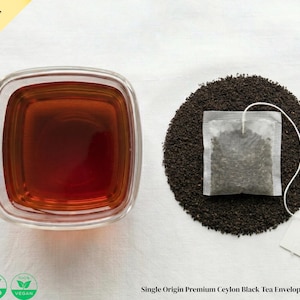 Puede incluir: Una taza cuadrada de vidrio llena de té oscuro junto a un lecho circular de hojas de té sueltas. Una bolsita de té con una etiqueta blanca se encuentra sobre las hojas. El texto de la imagen dice "CEYLON GREEN LAB" y "Single Origin Premium Ceylon Black Tea Enveloped Tea Bags".