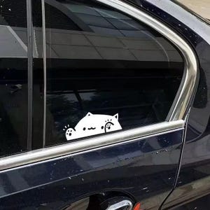 May include: A white vinyl decal applied to the dark tinted glass of a car window. The decal features a stylized graphic of a cat with pointed ears, a simple curved mouth, and two raised paws with visible paw pads, creating a playful appearance.