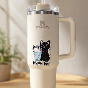 May include: A cream-colored Stanley tumbler with a handle, a clear lid, and a white straw. A sticker on the front features a black cat with colorful speckles drinking from a glass of water with the text Stay Hydrated in black lettering.