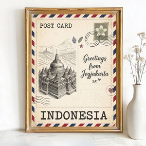 May include: Framed vintage postcard print with a detailed illustration of a temple in Yogyakarta, Indonesia. The postcard has a cream background with red, white, and blue accents, and the text "Greetings from Yogyakarta" and "Indonesia". The frame is gold.