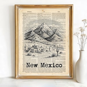 May include: Framed print featuring a black and white illustration of a desert landscape with mountains, cacti, and buildings. The artwork is set against a vintage dictionary page background, with the words "New Mexico" printed at the bottom. The frame is gold.