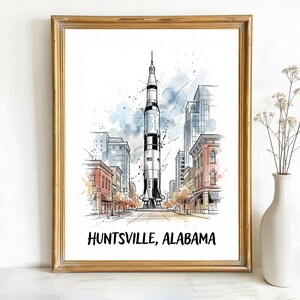 May include: Watercolour print of Huntsville, Alabama, depicting a rocket in a city street. The artwork is framed in a gold-coloured frame and includes the text "HUNTSVILLE, ALABAMA" at the bottom.