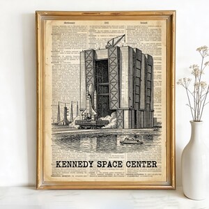 May include: A framed vintage print showing a black and white illustration of the Kennedy Space Centre. The artwork depicts a space shuttle on a launchpad, with the text "KENNEDY SPACE CENTER" at the bottom. The print is set against a backdrop of aged newspaper.
