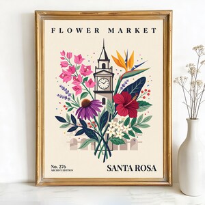 May include: A framed art print featuring a floral design with a clock tower. The print includes the text "FLOWER MARKET" and "SANTA ROSA". The artwork showcases pink, purple, red, and white flowers with green foliage against a cream background.