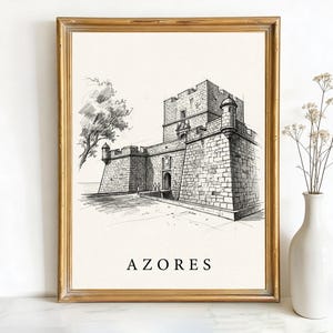 May include: A framed black and white sketch of a stone castle with the word "AZORES" in a serif font at the bottom. The artwork is in a gold frame and is next to a white vase with dried flowers.