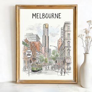 May include: Framed watercolor print of Melbourne, Australia, featuring a street scene with a green tram, pedestrians, and buildings. The word "MELBOURNE" is at the top. The print is in a gold frame, with a white vase and flowers to the right.