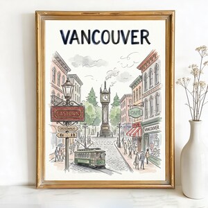 May include: Framed watercolor print of Vancouver, featuring a street scene with a green trolley, clock tower, and buildings. The word "Vancouver" is at the top. A white vase with dried flowers is to the right.