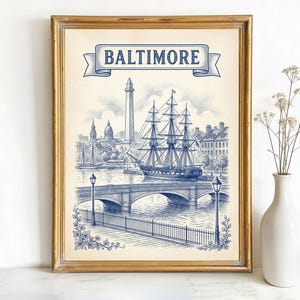 May include: Framed print featuring a blue and white illustration of Baltimore's harbor, with a tall ship, bridge, and buildings. The word "BALTIMORE" is displayed on a banner at the top. The frame is gold.
