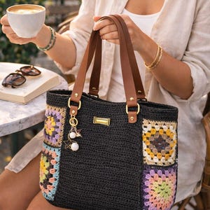 Puede incluir: Bolso tote de crochet negro con asas de cuero marrón y cuadrados de crochet coloridos. El bolso presenta herrajes dorados y un charm con estrellas y perlas. Un accesorio elegante y práctico para el uso diario.