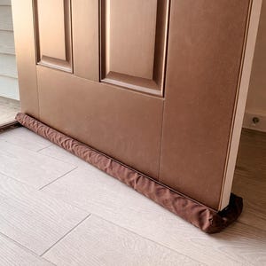 May include: A brown door with a matching brown draft stopper at the base. The door has a rectangular panel design. The draft stopper is a long, cylindrical shape, made of a soft, brown fabric. The floor is light-colored wood.