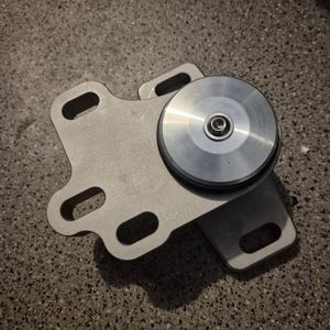 May include: A silver-colored metal mounting bracket featuring an irregular shape with multiple elongated slots for attachment. A circular, polished silver disc with a central bolt is securely fastened to the top of the bracket base.