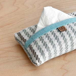 Peut inclure: Un porte-mouchoirs en tissu avec un motif en chevrons gris et blanc. Le bord supérieur est bordé d'une bande bleu sarcelle. Une petite étiquette en cuir marron est attachée. Des mouchoirs blancs sont visibles.