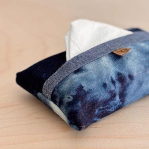 Peut inclure: Un étui à mouchoirs rectangulaire en tissu denim. Il présente un motif tie-dye bleu et blanc, avec une bordure bleu foncé et une petite étiquette en cuir. Des mouchoirs blancs sont visibles.
