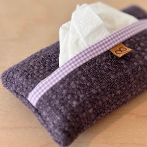 Peut inclure: Un porte-mouchoirs rectangulaire violet foncé en tissu texturé. Un ruban vichy blanc et lilas orne l'ouverture, révélant des mouchoirs blancs. Une petite étiquette en cuir marron avec un logo est attachée.