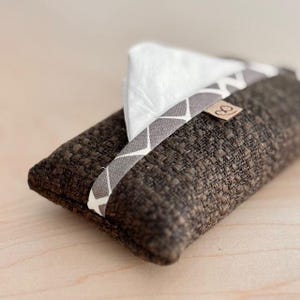 Peut inclure: Un étui à mouchoirs rectangulaire en tissu marron avec une bande à motifs géométriques gris et blancs. Des mouchoirs blancs sont visibles. Une petite étiquette marron est attachée à la bande.