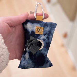 May include: A tie-dye blue and white dog waste bag dispenser with a silver carabiner clip and a brown leather accent. The dispenser has a metal grommet for easy bag access and a small brown leather patch with a logo.