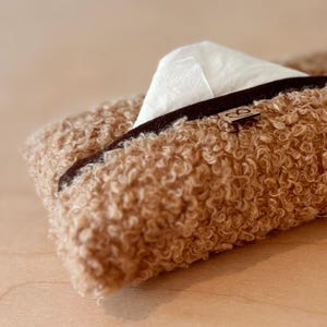 May include: A close-up of a brown, textured tissue holder with a dark brown trim. White tissues are partially visible, emerging from the opening. The holder has a soft, curly texture, and a small, decorative tag is attached.