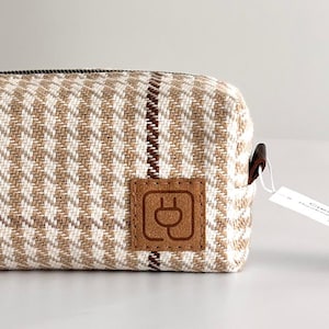 May include: A rectangular wash bag with a beige and white houndstooth pattern. It has a brown leather patch with a plug symbol and a brown leather handle. A brown zip and a tag are also visible.
