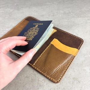 May include: A brown leather passport holder with a yellow pocket and a passport from Canada. The passport holder is open, revealing the passport inside. The holder is made of textured leather and has a stitched border.