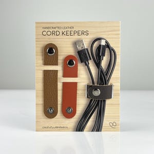 May include: Handcrafted leather cord keepers in brown and red, with a black USB cable. The card reads "CORD KEEPERS" and "creativityunlimited.ca". These cord keepers are designed to organise and secure cords.