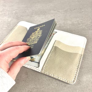 May include: A white and beige leather passport holder with a Canadian passport inside. The passport holder has multiple pockets and is open, revealing the passport and other cards. The holder is made of stitched leather.