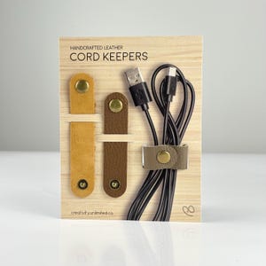 May include: A product display featuring handcrafted leather cord keepers in various shades of brown and tan, secured with gold-colored snaps. The display includes a bundle of black cords held together by a matching leather cord keeper. The text "CORD KEEPERS" is visible.