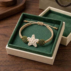 May include: A beige macrame bracelet with a white star and green leaf accents, presented in a green-lined box. The bracelet features gold-colored beads and is adjustable. The box is off-white with a gold-colored trim.
