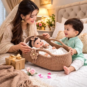 Könnte beinhalten: Ein brauner gehäkelter Babykorb mit einer Puppe darin, gehalten von einer Frau und einem Kind. Die Puppe trägt eine Schleife und liegt auf einer weißen Decke. Eine Geschenkbox und eine Decke befinden sich in der Nähe.