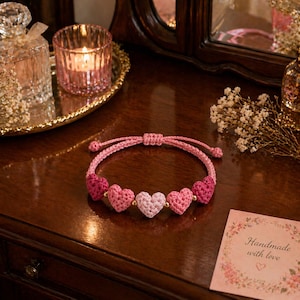 May include: A pink crochet bracelet featuring five heart-shaped charms in varying shades of pink. The bracelet is displayed on a dark wooden surface, with a small card that reads "Handmade with love".