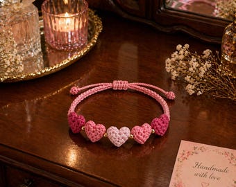 May include: A pink crochet bracelet featuring five heart-shaped charms in varying shades of pink. The bracelet is displayed on a dark wooden surface, with a small card that reads "Handmade with love".