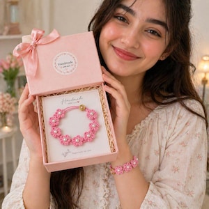 Puede incluir: Una caja de regalo rosa con un lazo a juego contiene una pulsera y una tarjeta hechas a mano. La pulsera presenta flores rosas de ganchillo y perlas de imitación. La tarjeta dice "Handmade with love" y "Especially for you". Se lleva una pulsera a juego.