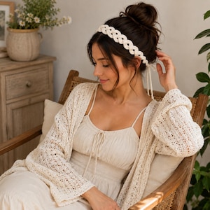May include: A cream-colored crocheted headband featuring an intricate braided pattern with thin ties at the back. The headband is worn with a matching cream-colored crocheted cardigan and a sleeveless top, both showcasing a delicate open-knit texture.