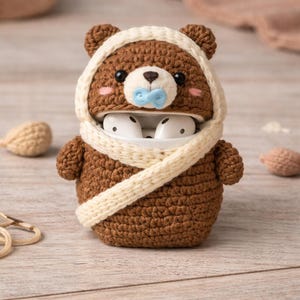 May include: A brown crocheted bear-shaped case for wireless earbuds. The bear has a white hood and scarf, pink cheeks, and a blue pacifier. The case is designed to hold white earbuds.