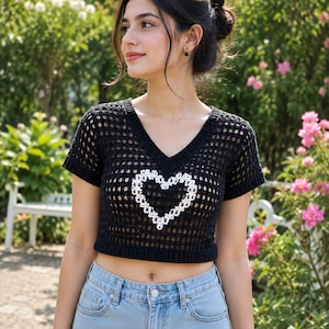 Può includere: Top corto all'uncinetto nero con un design a cuore bianco. Il top ha uno scollo a V e maniche corte. Il top è abbinato a jeans azzurri. Lo sfondo presenta un giardino con fiori rosa e verde.