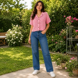 May include: A short-sleeved pink crochet button-down shirt with a collar, paired with blue denim jeans and white sneakers. The shirt features a textured, open-knit pattern and a row of buttons down the center, while the jeans have a straight-leg fit.
