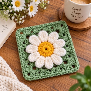 May include: A square crochet coaster featuring a sage green granny square base with a three-dimensional white daisy flower with a bright yellow center stitched onto the middle. The yarn texture is visible throughout the handmade square piece.
