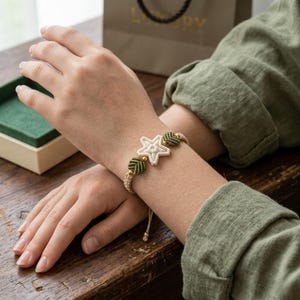 May include: A macrame bracelet featuring a cream-colored star and green leaf accents. The bracelet is made with beige cord and gold-colored beads. The bracelet is on a person's wrist.