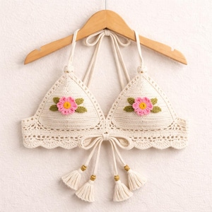 Puede incluir: Top de crochet color crema con cuello halter y detalles florales. El top presenta un diseño triangular con aplicaciones de flores rosas y verdes en cada copa. Borlas con cuentas doradas cuelgan de la parte inferior, añadiendo un toque bohemio.