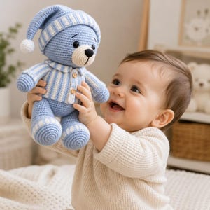 Puede incluir: Un oso de peluche tejido a ganchillo en azul claro y blanco. El oso lleva un gorro de dormir a rayas a juego con un pompón blanco, una parte superior de pijama a rayas con dos pequeños botones de madera y puños a rayas en los pies. Tiene un hocico color crema.