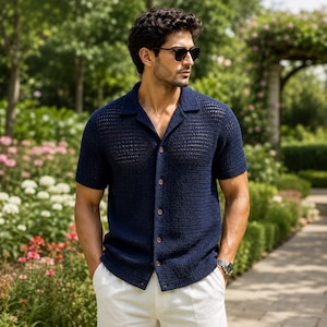 May include: A navy blue short-sleeved button-down shirt featuring a textured, open-knit crochet pattern with a classic collar. The shirt is paired with plain white trousers, creating a clean, structured look with visible button detailing.