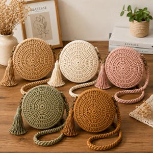 May include: Five round crochet crossbody bags in tan, cream, dusty pink, sage green, and mustard yellow. Each bag features a circular textured pattern, a matching long shoulder strap, and a decorative tassel attached to the side with bronze hardware.