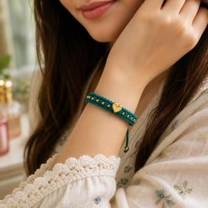 May include: A teal macrame bracelet with gold-coloured beads and a heart charm. The bracelet is worn on a wrist, showcasing the intricate knotting and the charm's placement. The bracelet's design is simple yet elegant.