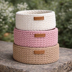 Peut inclure: Trois paniers ronds en crochet empilés, de couleur crème, rose et beige. Chaque panier porte une étiquette en cuir avec le mot "MONOMEY". Les paniers sont faits de fil épais et sont posés sur une surface en pierre.