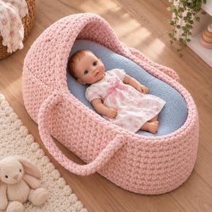 May include: A baby doll with light brown hair and blue eyes wearing a white floral dress with a pink bow, lying inside a soft, hand-crocheted pink bassinet with a light blue interior lining and two sturdy handles on the sides.